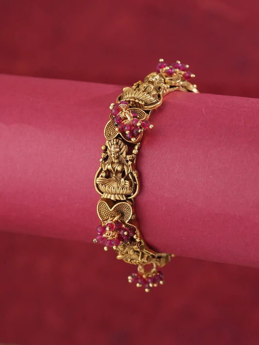An ornate gold-toned bracelet featuring detailed filigree and small red gemstone accents, displayed on a pink cylindrical stand against a matching red background.