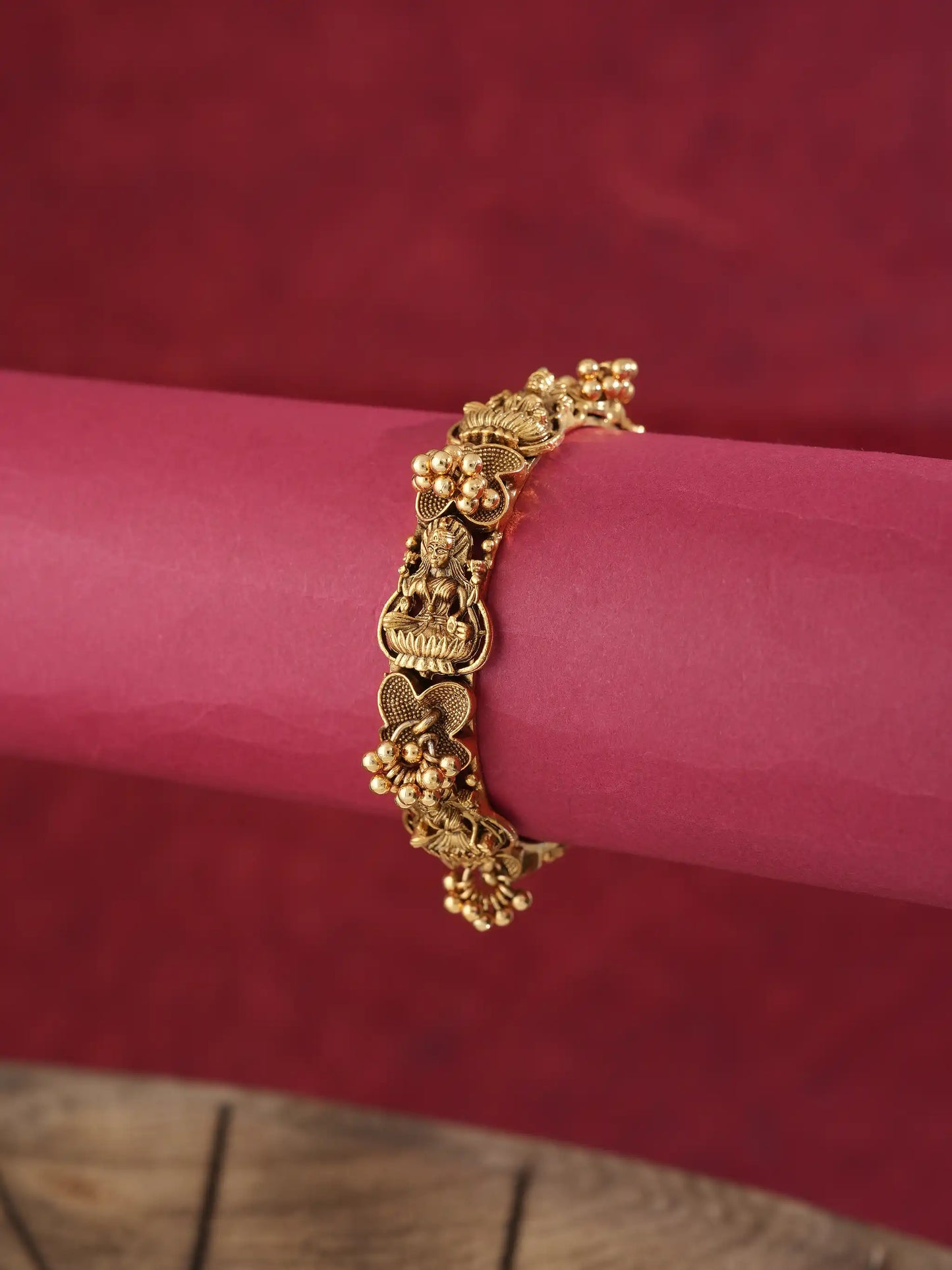 A gold-toned traditional Indian bracelet with detailed motifs and small bead clusters, displayed on a pink cylindrical stand against a dark red background.