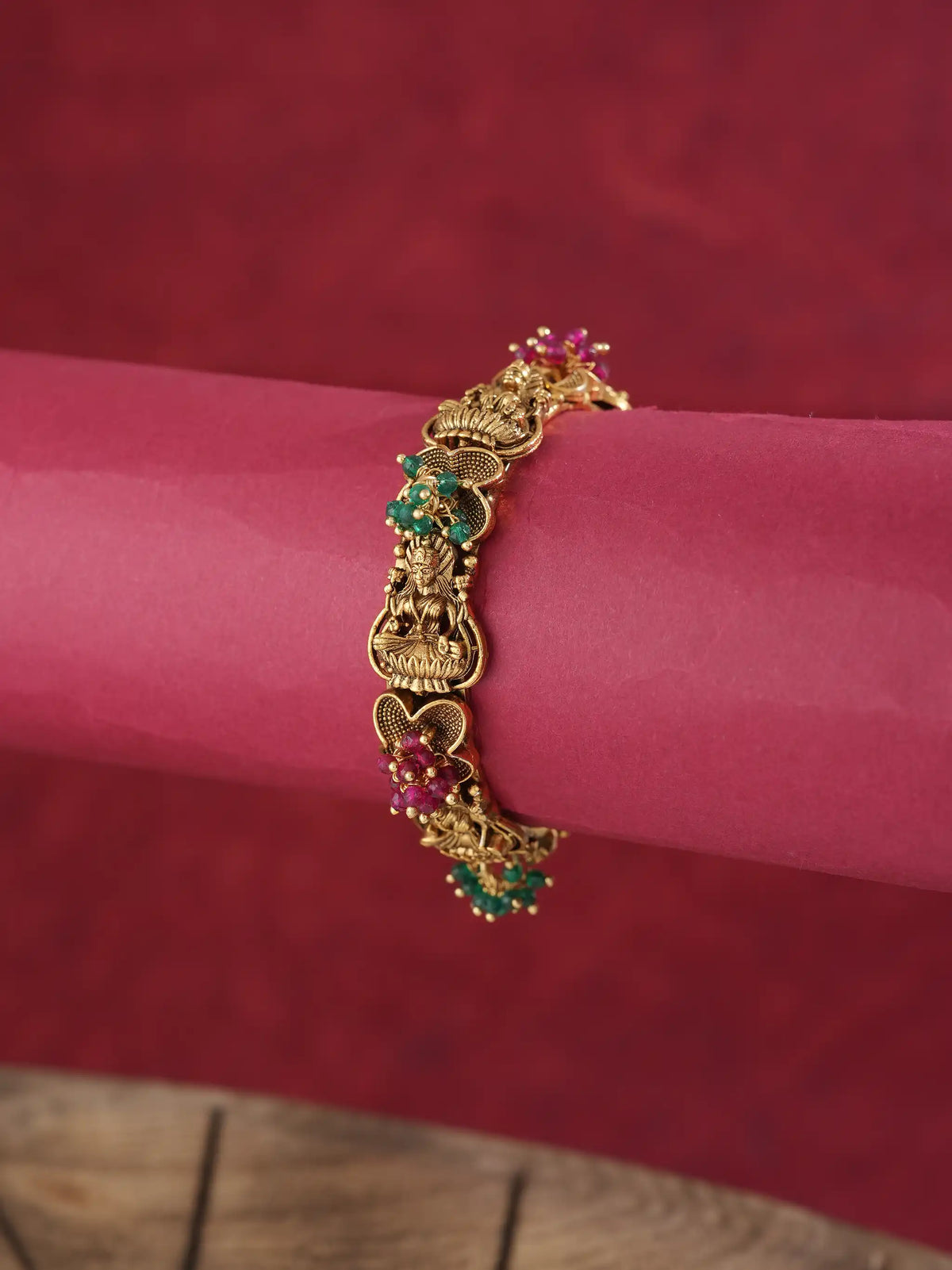 An ornate gold-toned bracelet with detailed motifs and small green and pink gemstone accents, shown worn on a pink cylindrical display against a dark red textured background.