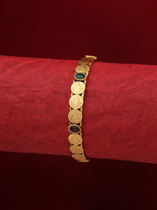 A gold-toned bracelet featuring multiple circular coin-like links with inset dark green and dark red stones, displayed on a textured red cylindrical stand against a dark red background.