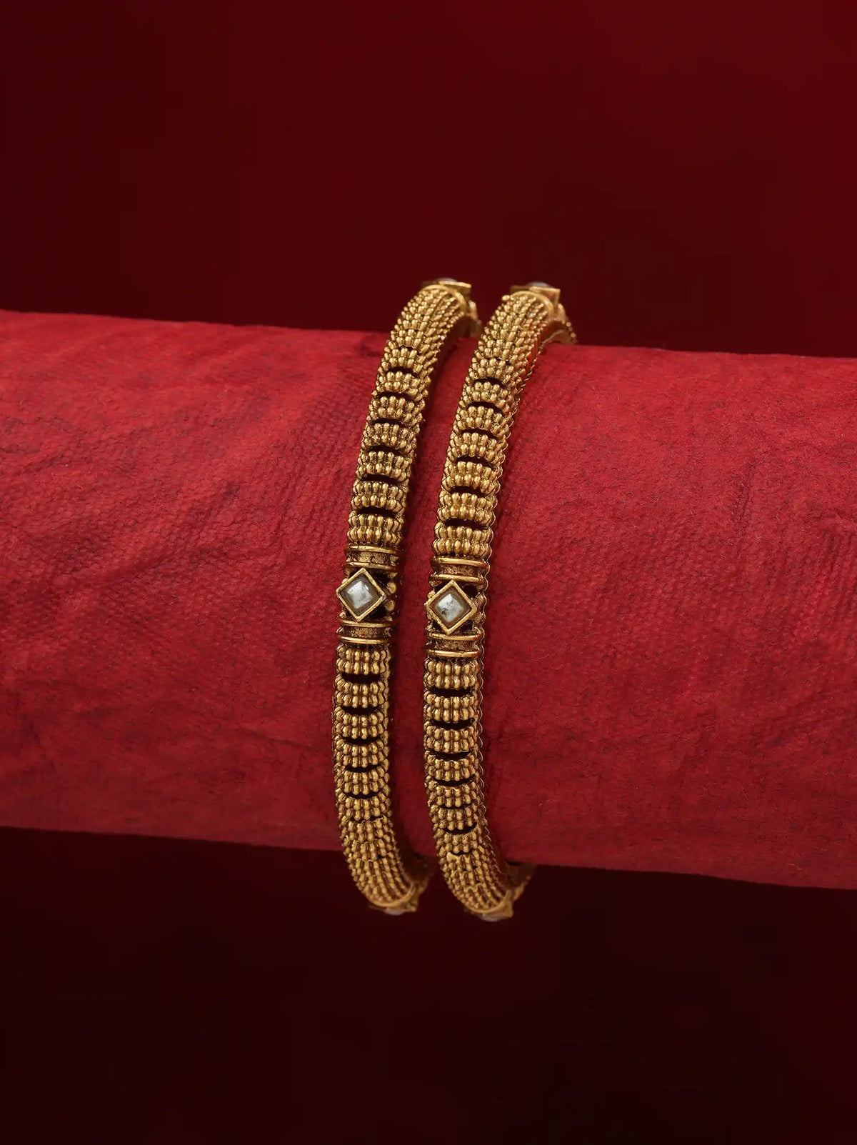 Two gold-toned beaded bangles with small diamond-shaped white inlays, displayed on a textured red cylindrical stand against a dark red background.