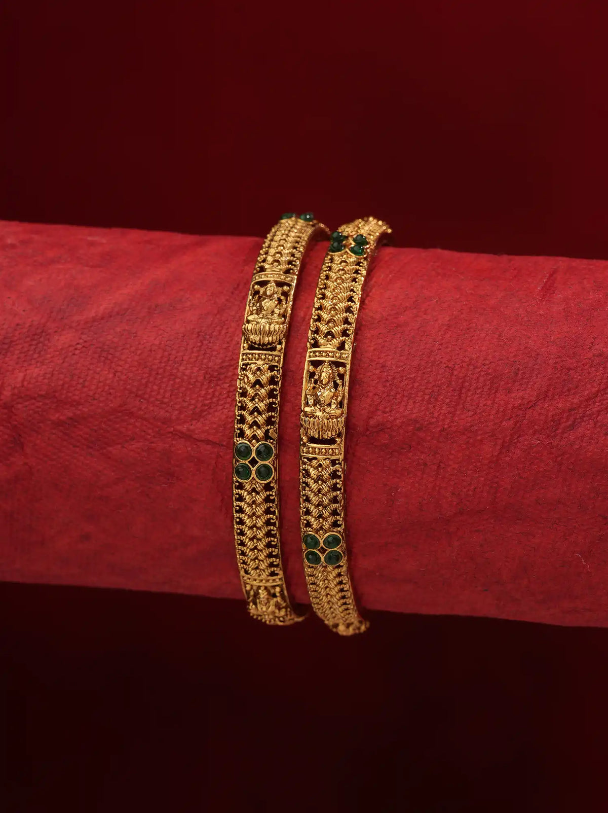 Two ornate gold bangles with green stone accents, displayed on a textured red cylindrical stand against a dark red background.