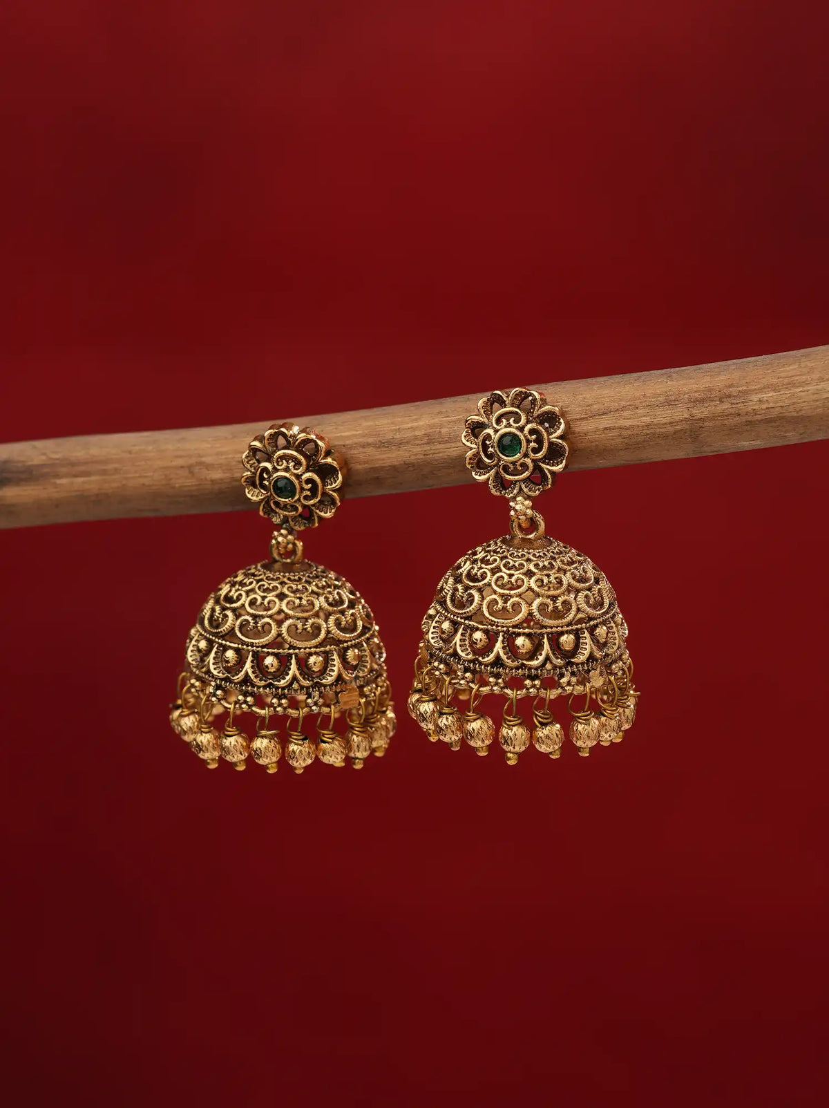 A pair of ornate gold-toned jhumka earrings with small green stone accents and dangling bead chains, hanging from a wooden branch against a solid dark red background.