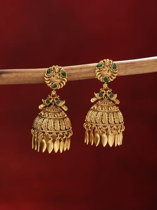 A pair of traditional Indian gold jhumka earrings with green stone accents, hanging from a wooden branch against a dark red background.