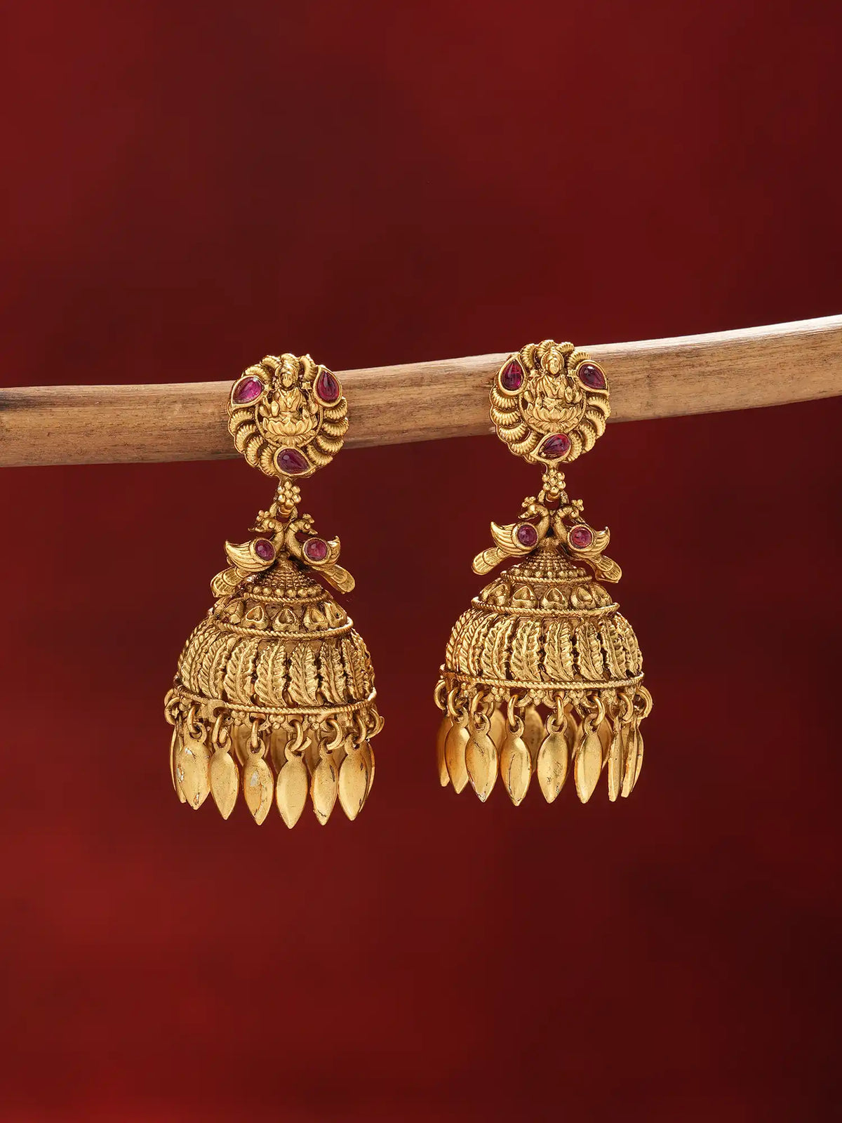 A pair of ornate gold-toned Indian jhumka earrings with small red gemstone accents, hanging from a wooden branch against a dark red background.