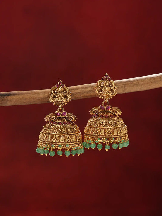 A pair of ornate gold-toned jhumka earrings with green bead fringe and small red stone accents, hanging from a wooden branch against a deep red background.
