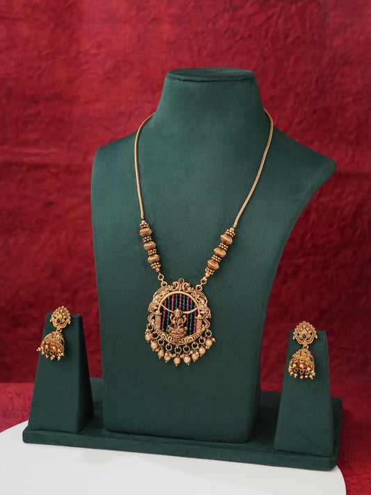Gold-toned traditional Indian jewelry set featuring a necklace and matching earrings displayed on a dark green velvet jewelry stand against a textured red backdrop.