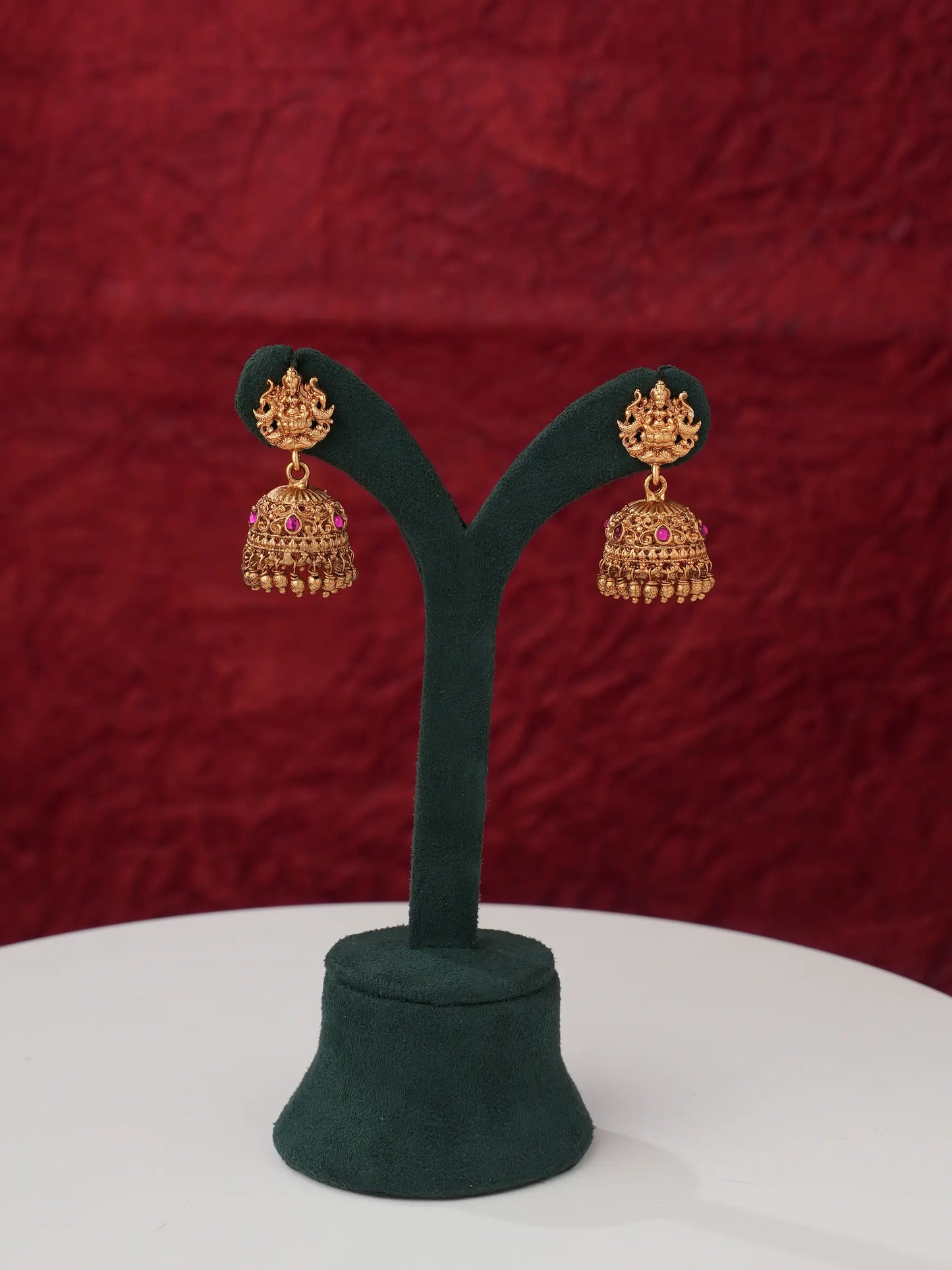 A pair of ornate gold-toned jhumka earrings with pink stone accents, displayed on a dark green Y-shaped jewelry stand against a textured dark red backdrop.