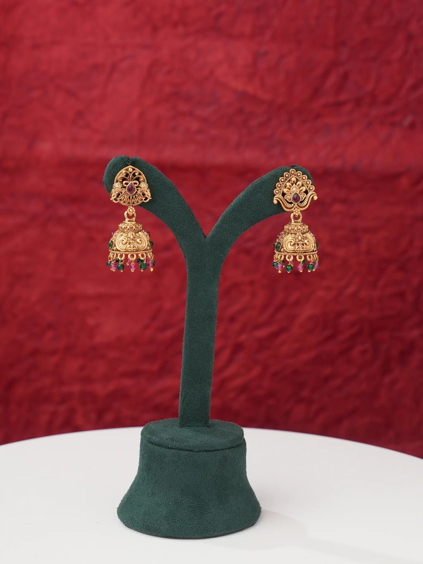 A pair of ornate gold-toned Indian jhumka earrings with red and green bead accents, displayed on a dark green velvet jewelry stand against a textured red background.