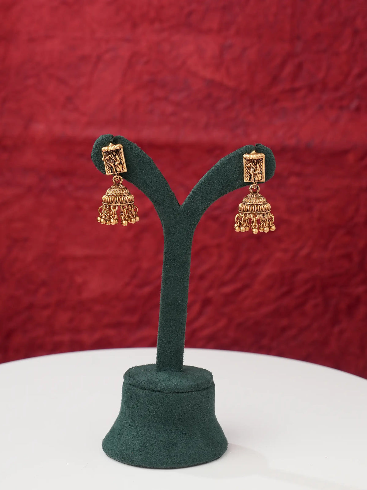 A pair of gold-toned traditional Indian jhumka earrings displayed on a dark green velvet jewelry stand against a textured red background.
