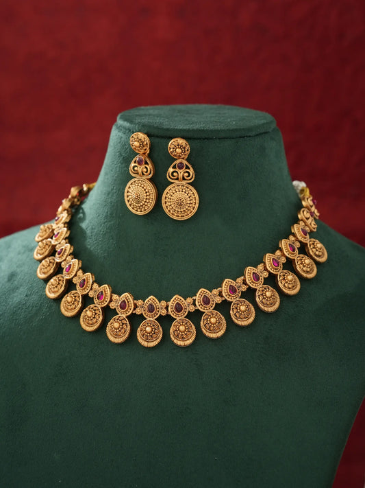 Gold-toned traditional jewelry set featuring a necklace and matching earrings displayed on a dark green velvet jewelry bust against a textured red background.