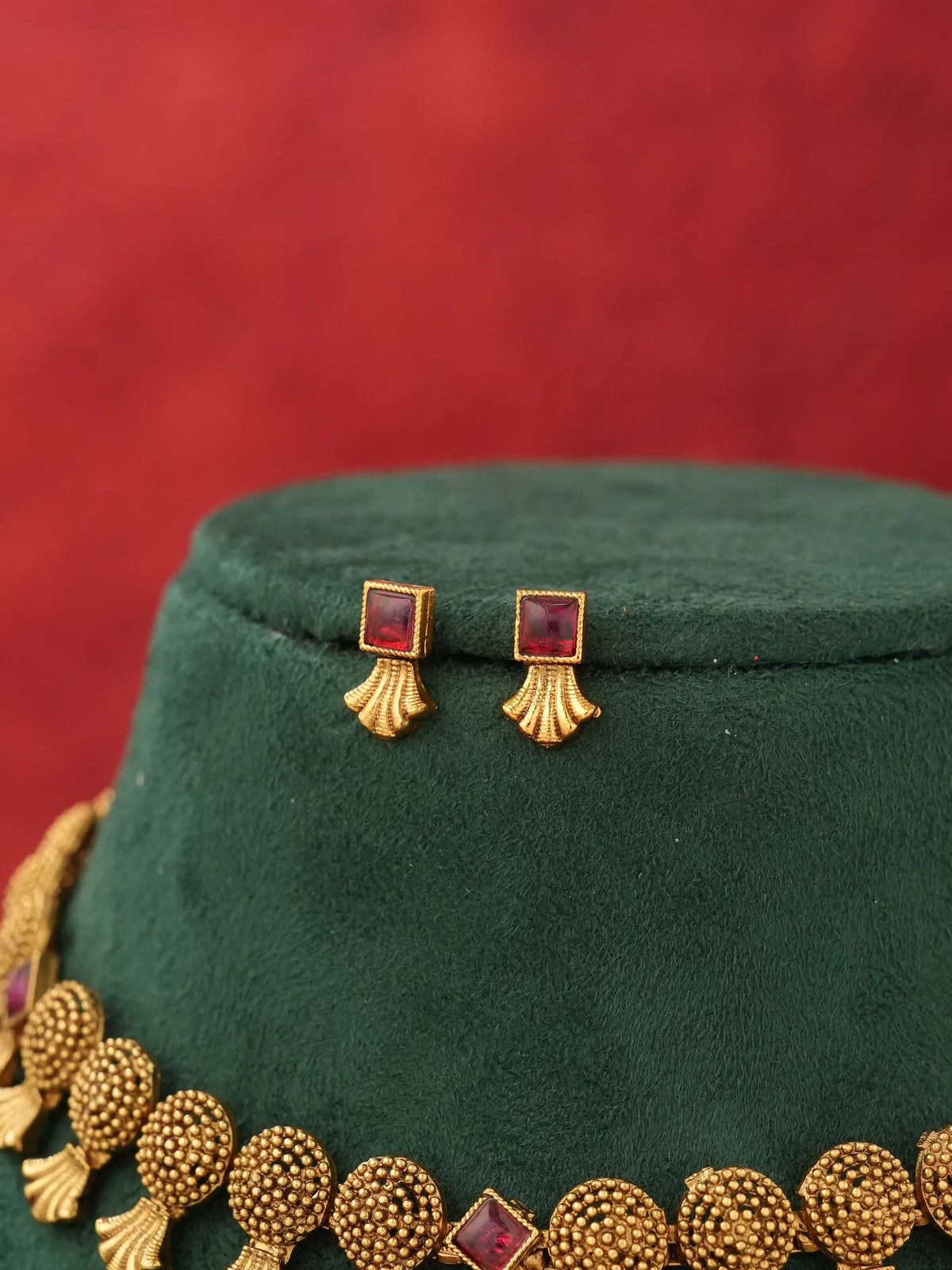 Gold-toned jewelry set featuring a necklace and matching earrings with red square stones and fan-shaped gold details, displayed on a dark green velvet stand against a red background.