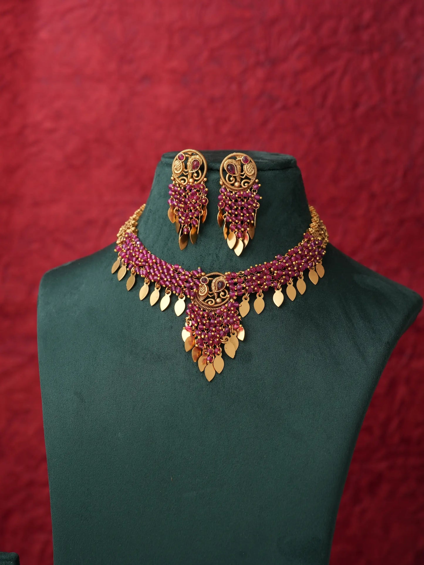 Gold and deep pinkish-red beaded jewelry set including a choker necklace and matching earrings, displayed on a dark green velvet jewelry bust against a textured red background.