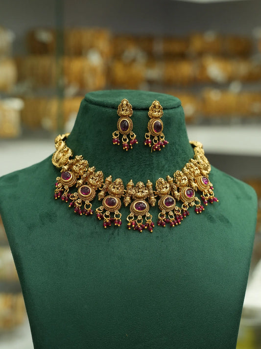 An ornate gold-toned traditional necklace and matching drop earrings displayed on a dark green velvet jewelry bust. The jewelry features circular purple stones and dangling red bead accents, with a softly blurred shop interior in the background.