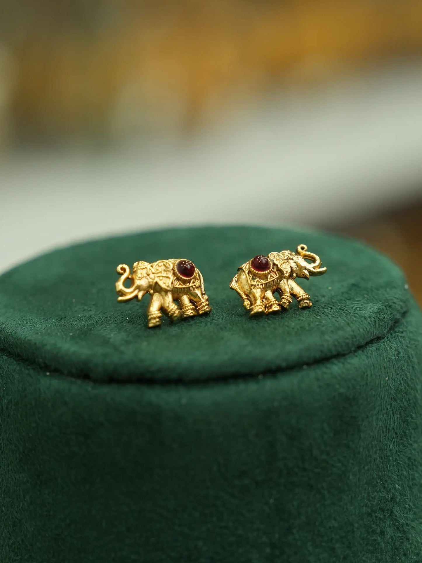 A pair of gold elephant stud earrings with small red gemstone accents, displayed on a dark green velvet jewelry stand.
