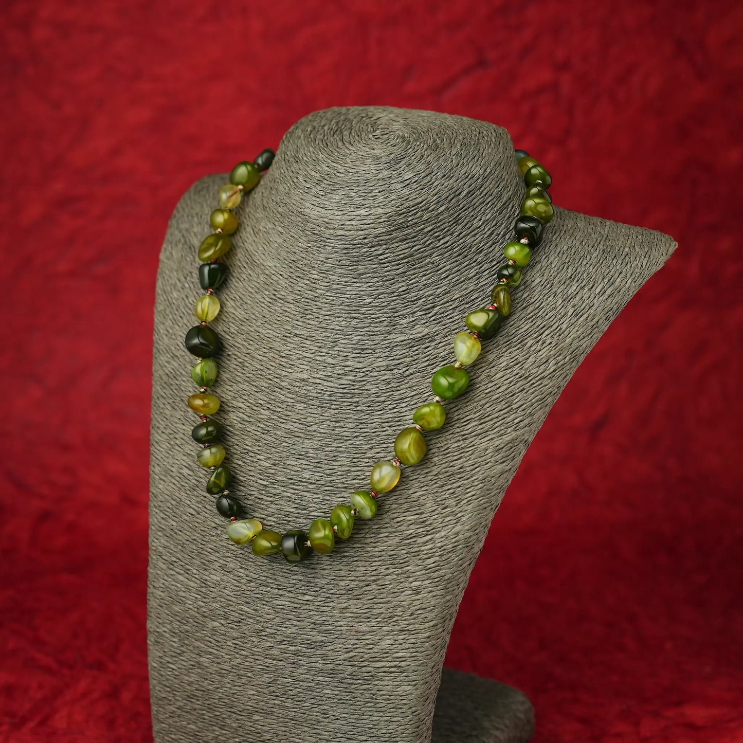 A beaded necklace made of polished olive green and dark forest green marbled beads, displayed on a gray fabric jewelry bust against a textured red background.
