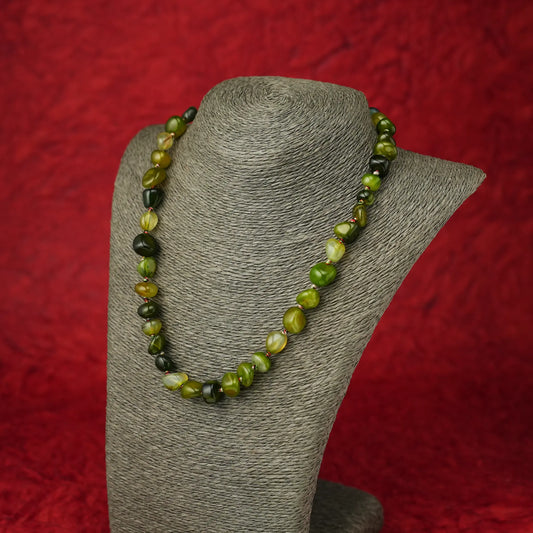 A beaded necklace made of polished olive green and dark forest green marbled beads, displayed on a gray fabric jewelry bust against a textured red background.