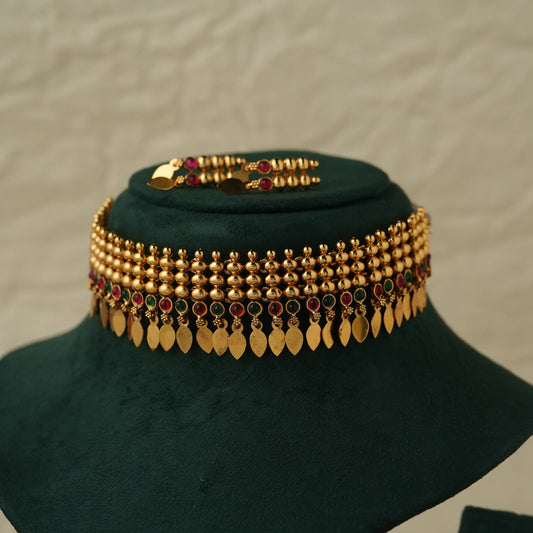 A gold-toned traditional necklace with beaded strands, red and green stone accents, and dangling gold leaf-shaped charms, displayed on a dark green velvet jewelry bust against a light textured background.