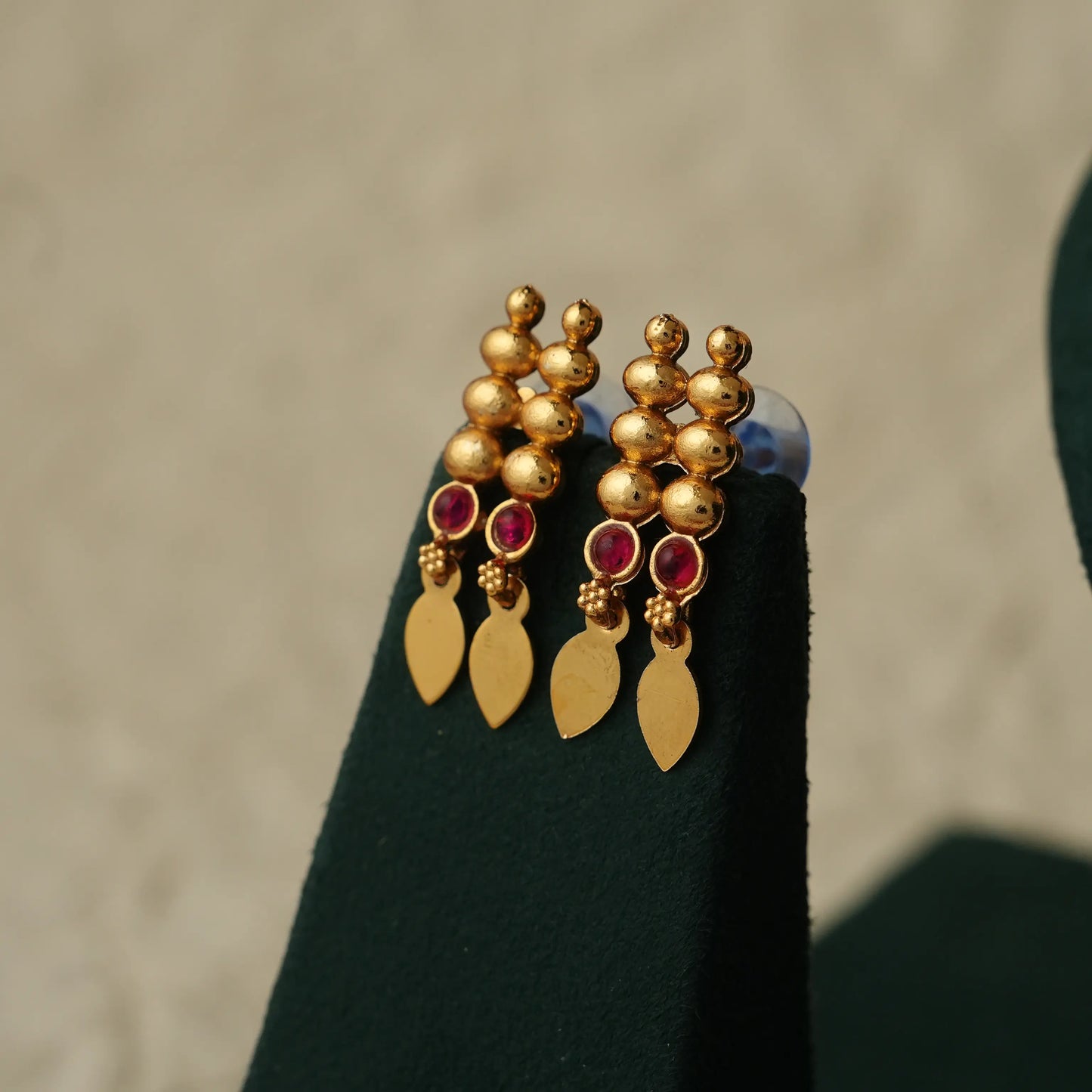 A pair of gold-toned dangle earrings with small red gemstone accents and teardrop-shaped gold charms, displayed on a black jewelry stand against a neutral beige background.