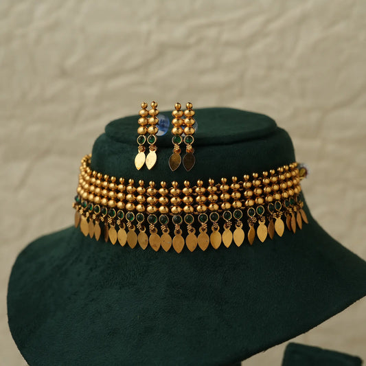 Gold-toned traditional jewelry set displayed on a dark green velvet jewelry bust, featuring a beaded necklace with hanging leaf-shaped coins, matching drop earrings, and a small blue stone accent.