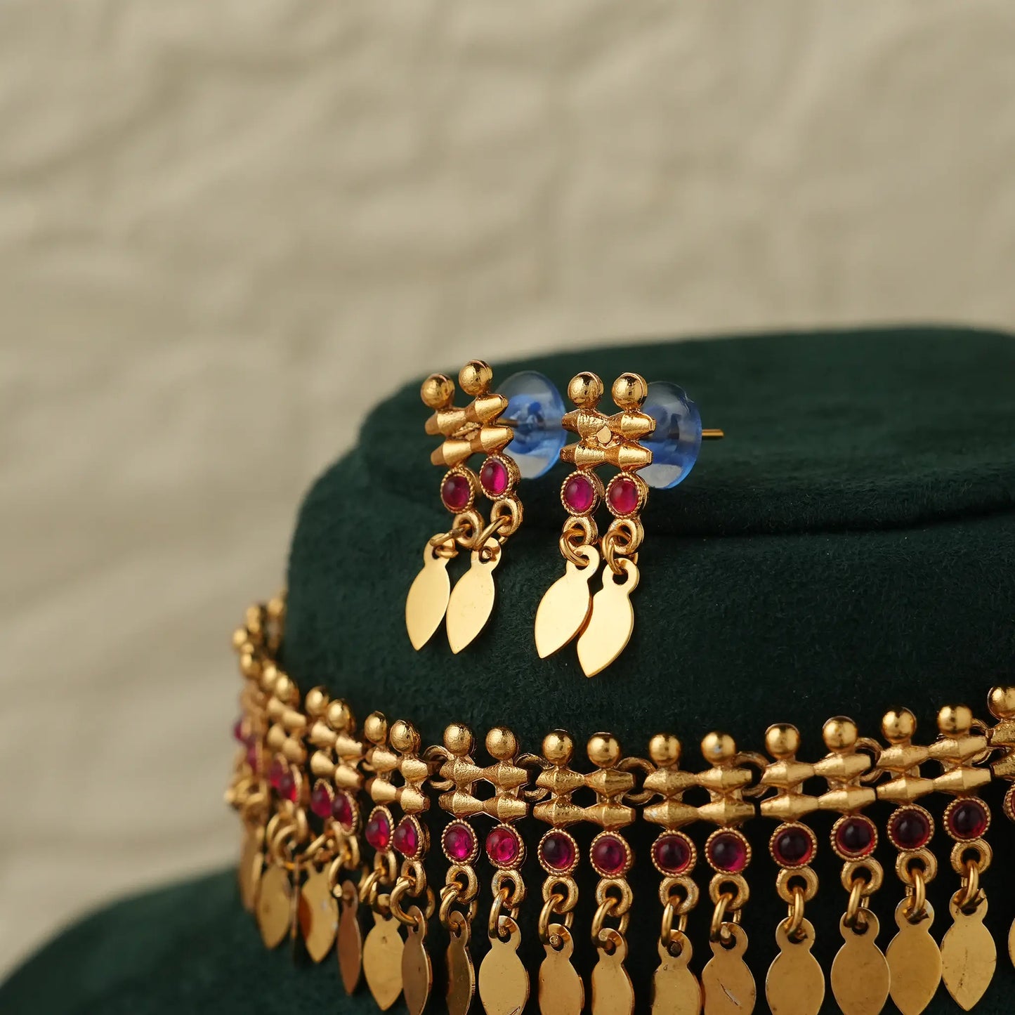 Gold-toned traditional jewelry set featuring a choker necklace and matching drop earrings, decorated with gold beads, and deep pink round stones, displayed on a dark green velvet stand.