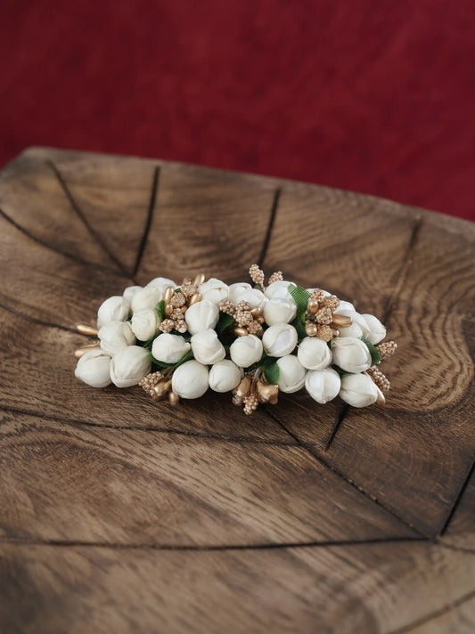 A white jasmine flower hair clip with gold-toned metal accents and small green leaves, displayed on a rustic wooden slice with a red textured background.