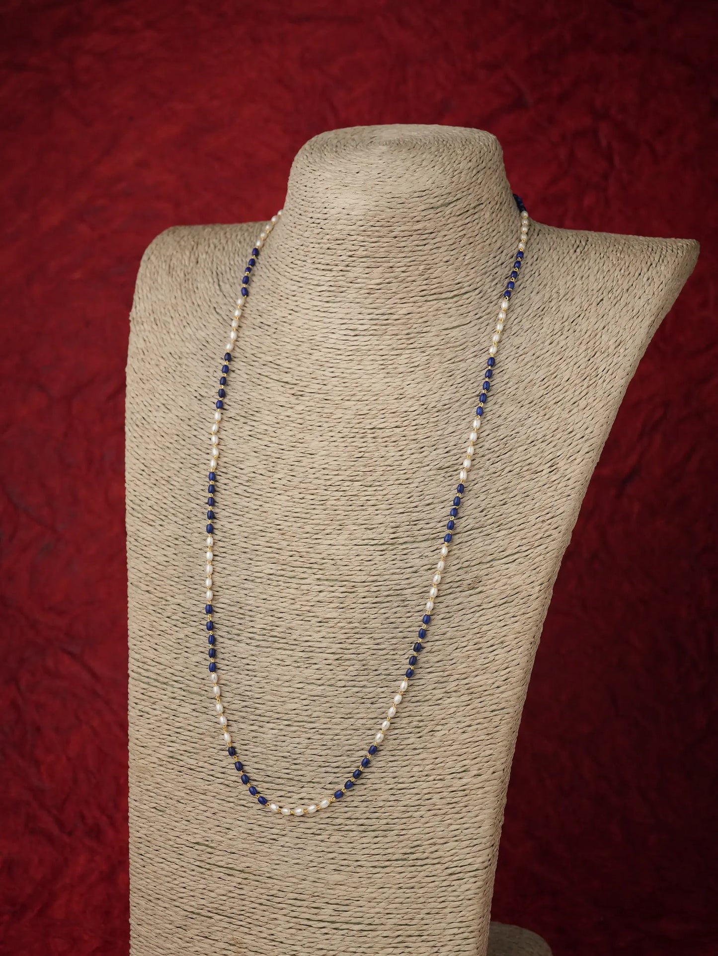 A long beaded necklace with alternating dark navy blue and cream beads, displayed on a beige fabric-covered jewelry bust against a textured dark red background.