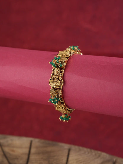 An ornate gold-toned bracelet with green stone accents, displayed on a rolled pink paper cylinder against a dark red textured background.