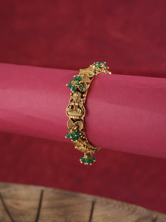 An ornate gold-toned bracelet with green stone accents, displayed on a rolled pink paper cylinder against a dark red textured background.