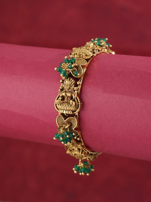 An ornate gold-toned bangle bracelet with detailed filigree, floral motifs, and green stone accents, displayed on a pink cylindrical stand against a dark red background.