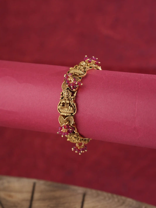 An ornate gold-toned bracelet with detailed filigree and small pink gemstone accents, displayed on a pink cylindrical stand against a textured red background.