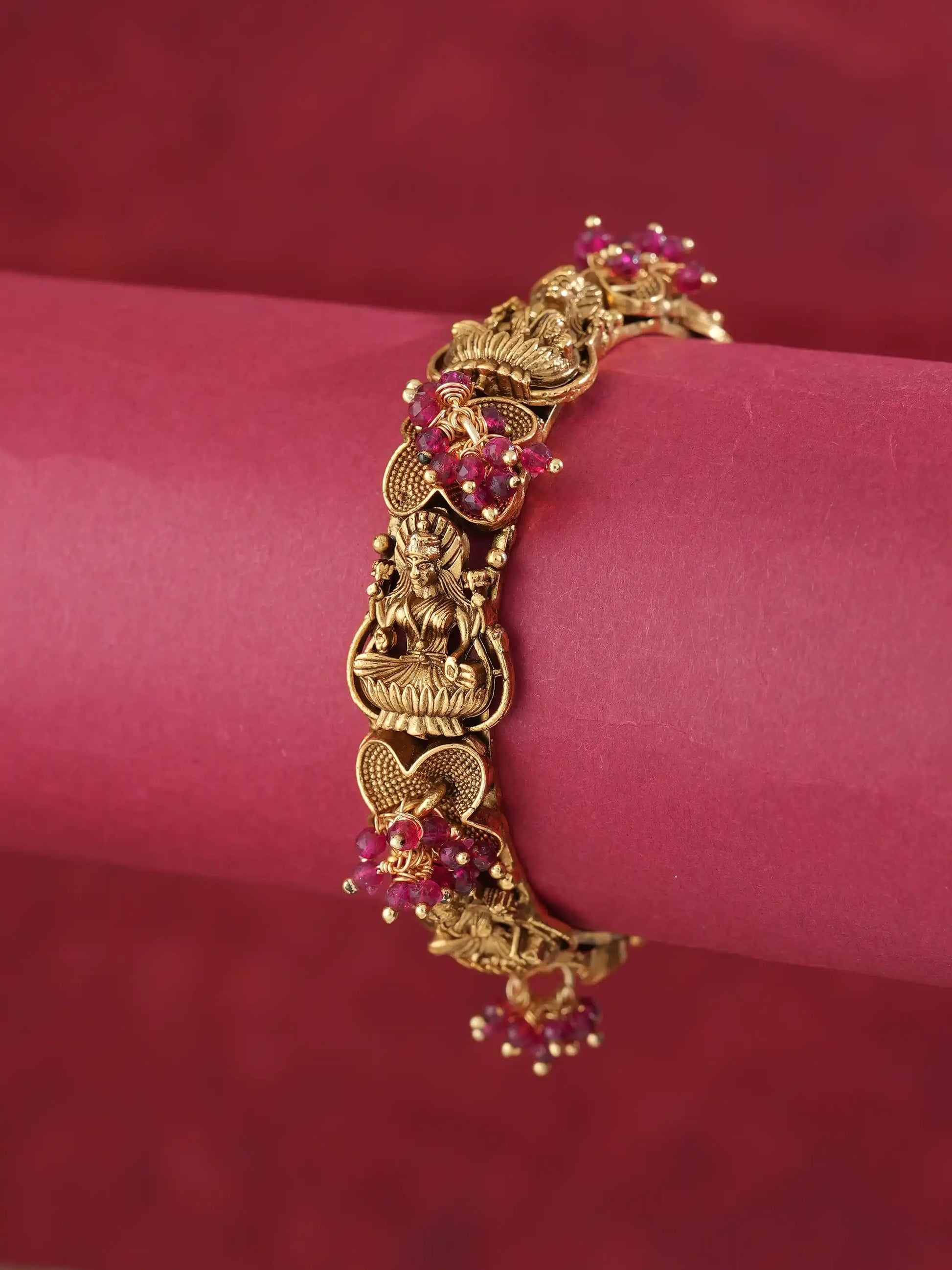 An ornate gold-toned bracelet featuring detailed filigree and small red gemstone accents, displayed on a pink cylindrical stand against a matching red background.