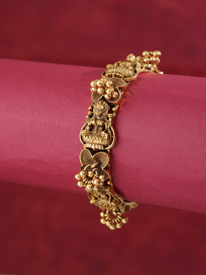 A gold-plated traditional Indian bracelet with detailed motifs of Lord Ganesha, lotus flowers, and small gold beads, displayed on a pink cylindrical stand against a dark red background.