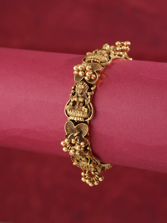 A gold-plated traditional Indian bracelet with detailed motifs of Lord Ganesha, lotus flowers, and small gold beads, displayed on a pink cylindrical stand against a dark red background.