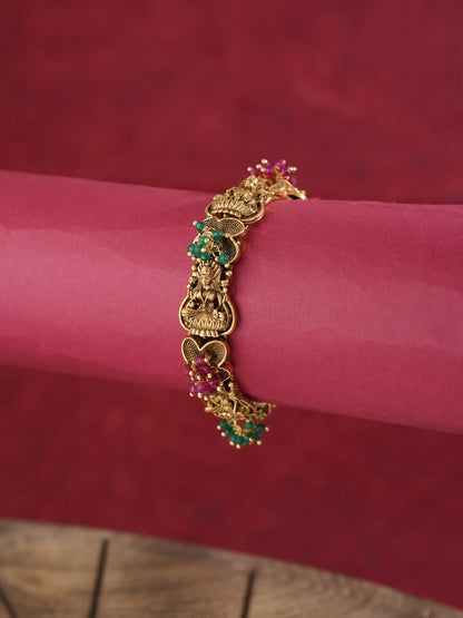 An ornate gold-toned bracelet with detailed motifs and small green and pink gemstone accents, shown worn on a pink cylindrical display against a dark red textured background.