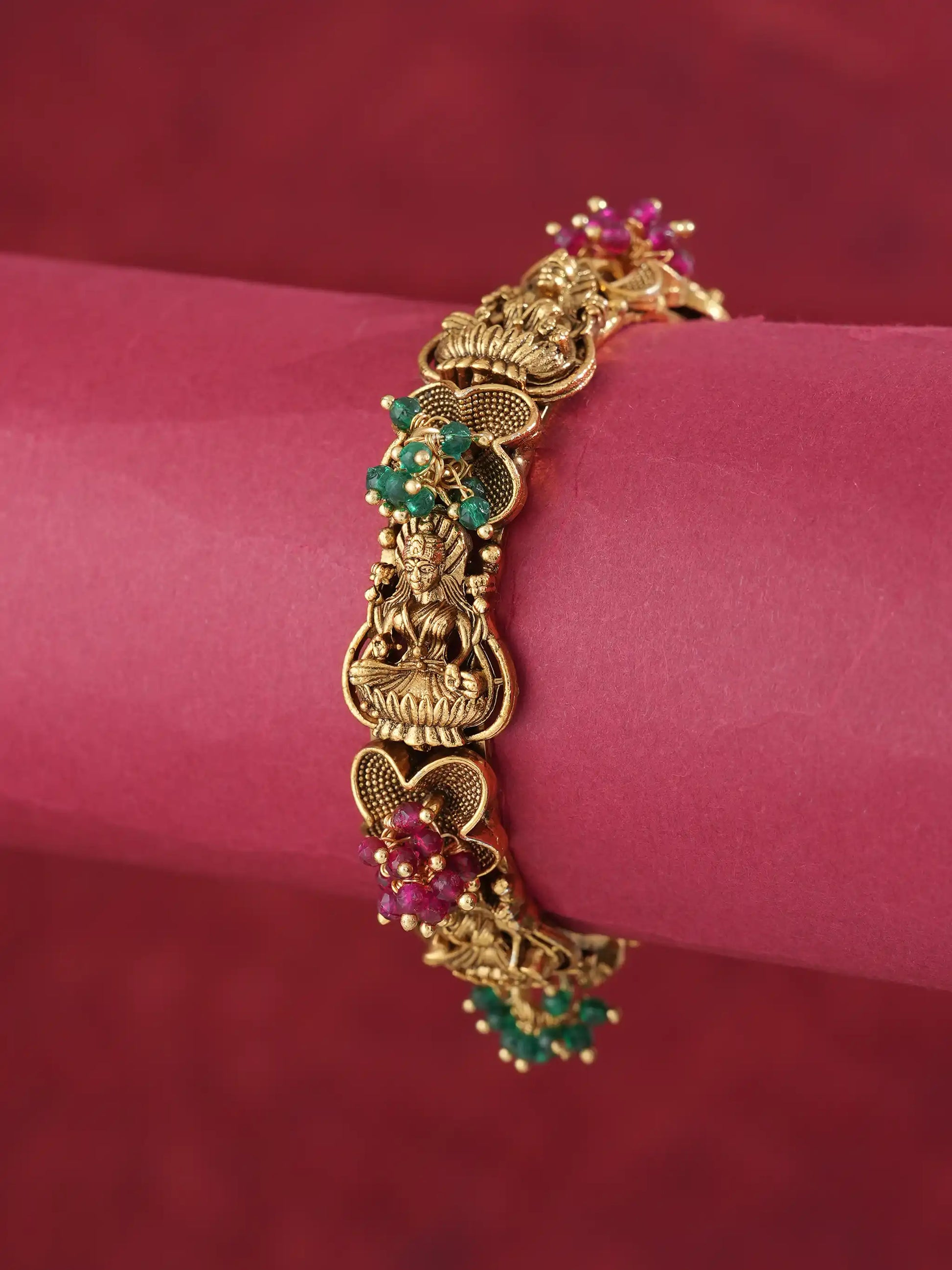 An ornate gold-toned bracelet with detailed filigree and small green and pink gemstone accents, displayed on a pink cylindrical stand against a dark red background.