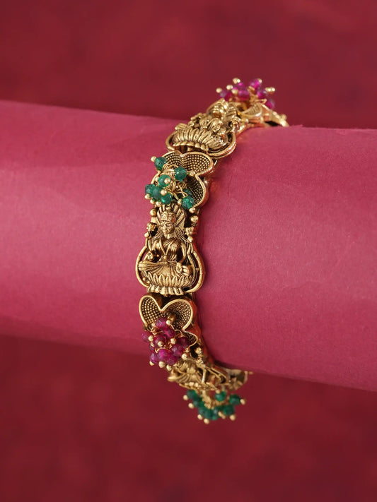 An ornate gold-toned bracelet with detailed filigree and small green and pink gemstone accents, displayed on a pink cylindrical stand against a dark red background.