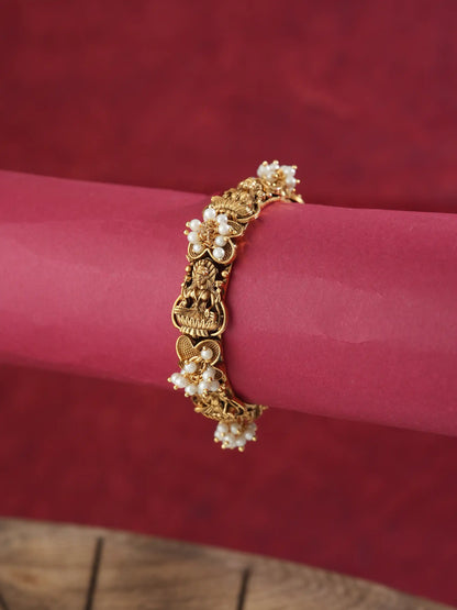 A gold-plated traditional Indian bracelet decorated with small white pearl flowers and a central deity charm, displayed on a pink cylindrical stand against a textured red background.