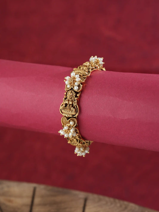 A gold-plated traditional Indian bracelet decorated with small white pearl flowers and a central deity charm, displayed on a pink cylindrical stand against a textured red background.