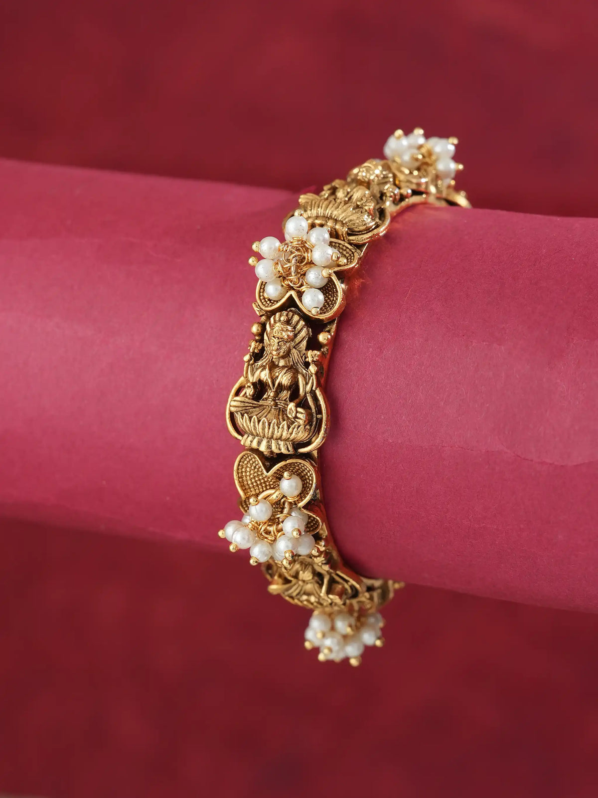An ornate gold-toned bracelet with floral pearl clusters and a central deity motif, displayed on a pink cylindrical stand against a dark red background.