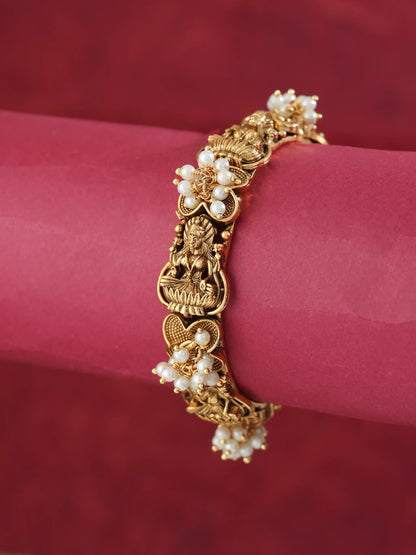 An ornate gold-toned bracelet with floral pearl clusters and a central deity motif, displayed on a pink cylindrical stand against a dark red background.