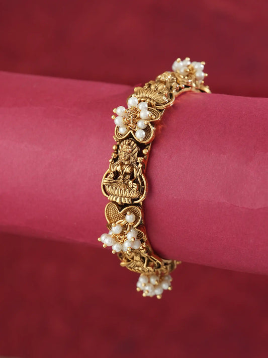 An ornate gold-toned bracelet with floral pearl clusters and a central deity motif, displayed on a pink cylindrical stand against a dark red background.