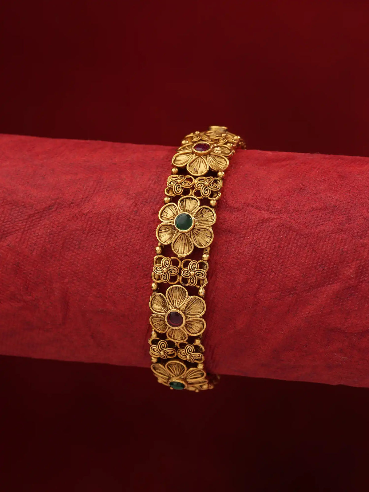 A gold-toned traditional Indian bangle bracelet with floral motifs and small green and red stone accents, displayed on a textured red fabric roll against a dark red background.