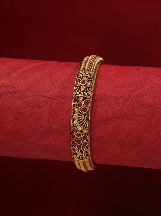 A gold-plated traditional Indian bangle bracelet with intricate filigree work and small red stone accents, displayed on a textured red cylindrical stand against a solid red background.