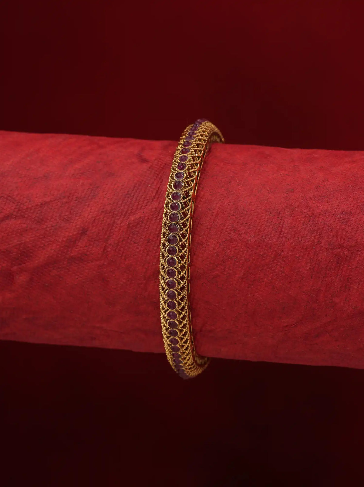 A gold-plated bangle bracelet with a row of dark red oval stones, displayed on a textured red cylindrical stand against a dark red background.