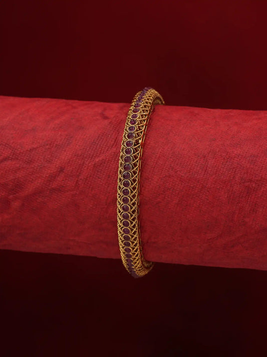 A gold-plated bangle bracelet with a row of dark red oval stones, displayed on a textured red cylindrical stand against a dark red background.