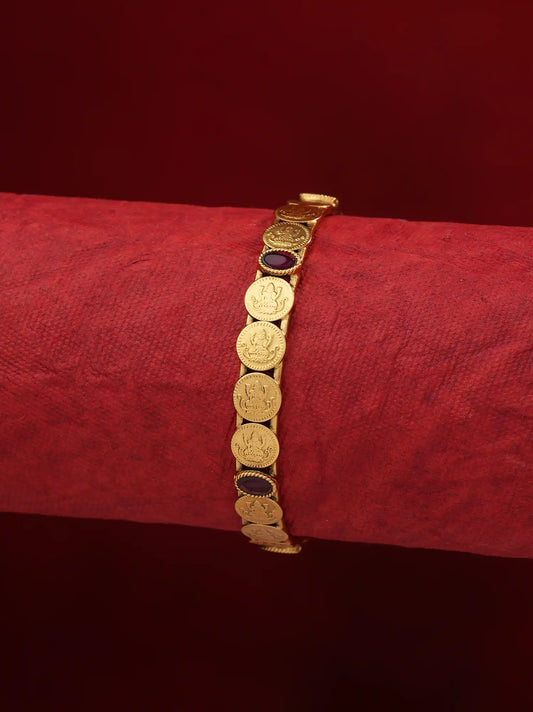 A gold-toned coin-link bracelet displayed on a textured red cylindrical stand against a dark red background.
