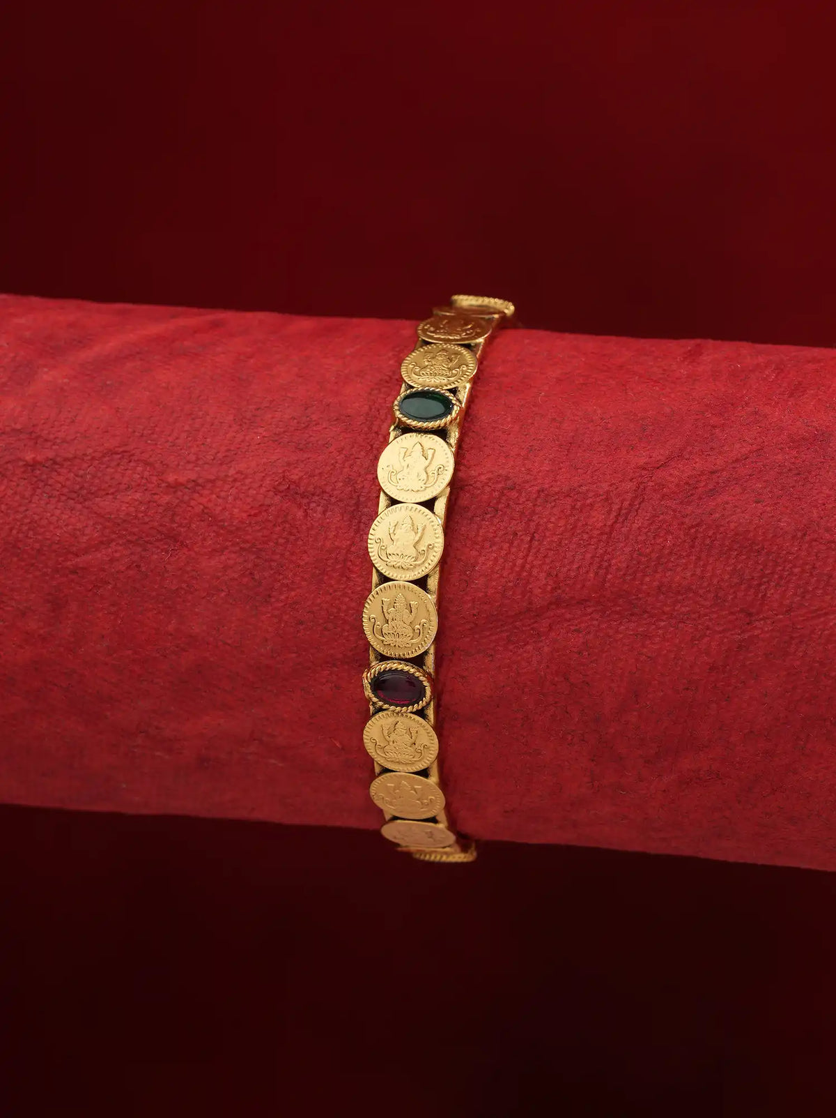 A gold-toned bracelet featuring multiple circular coin-like links with inset dark green and dark red stones, displayed on a textured red cylindrical stand against a dark red background.