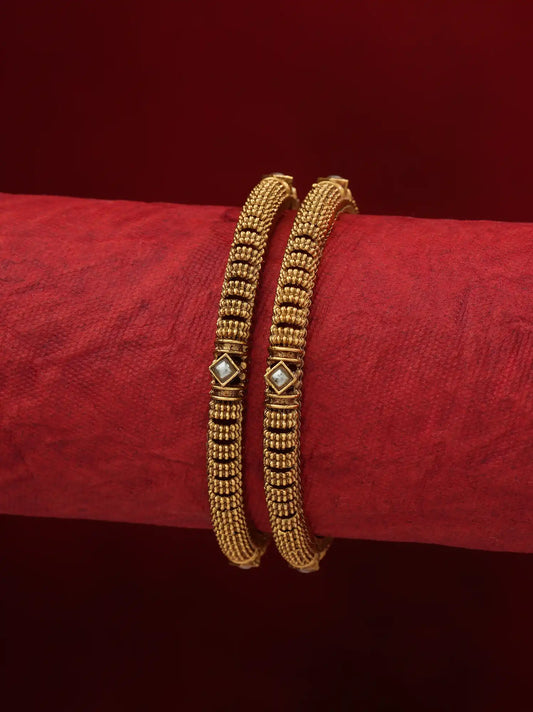 Two gold-toned beaded bangles with small diamond-shaped white inlays, displayed on a textured red cylindrical stand against a dark red background.