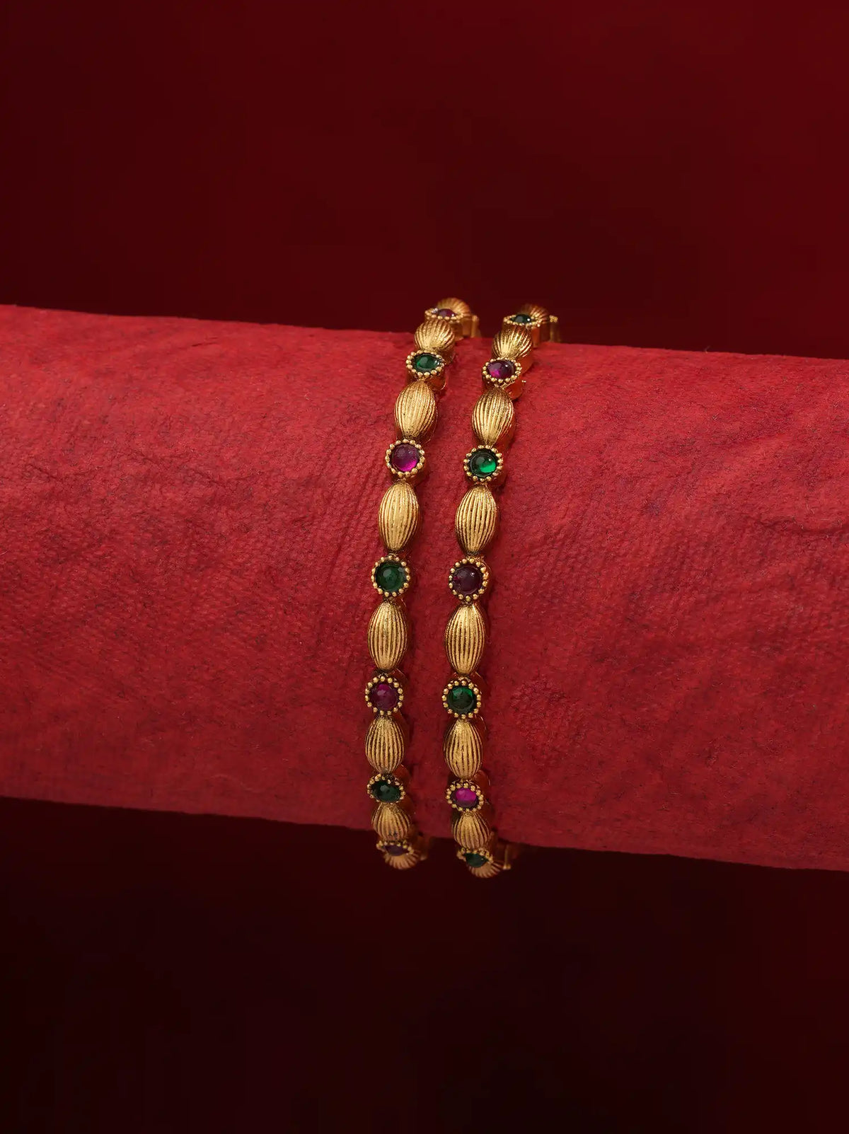 A pair of gold-plated bangles decorated with alternating oval textured beads and round green and pinkish-red gemstone accents, displayed on a textured red fabric roll against a dark red background.